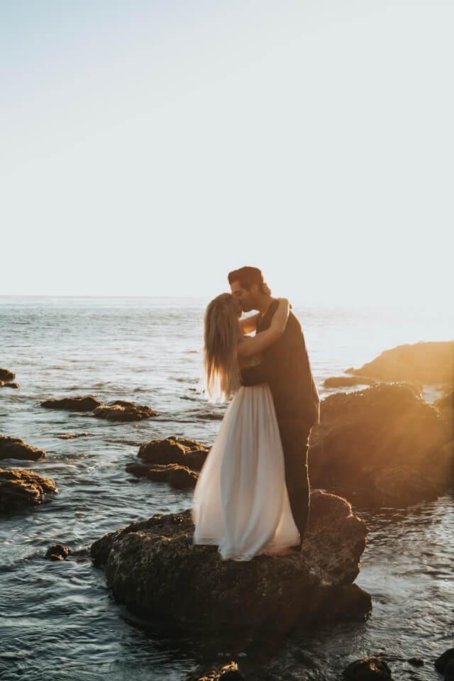 bride and groom kissing on rocks at sunset by the ocean